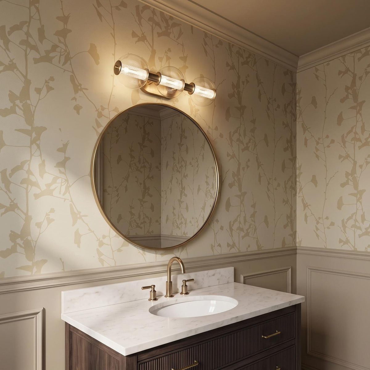 A transitional bathroom featuring the Elara LED 3 Light Vanity 25 in brass and clear glass above a round mirror, marble countertop, and botanical wallpaper.