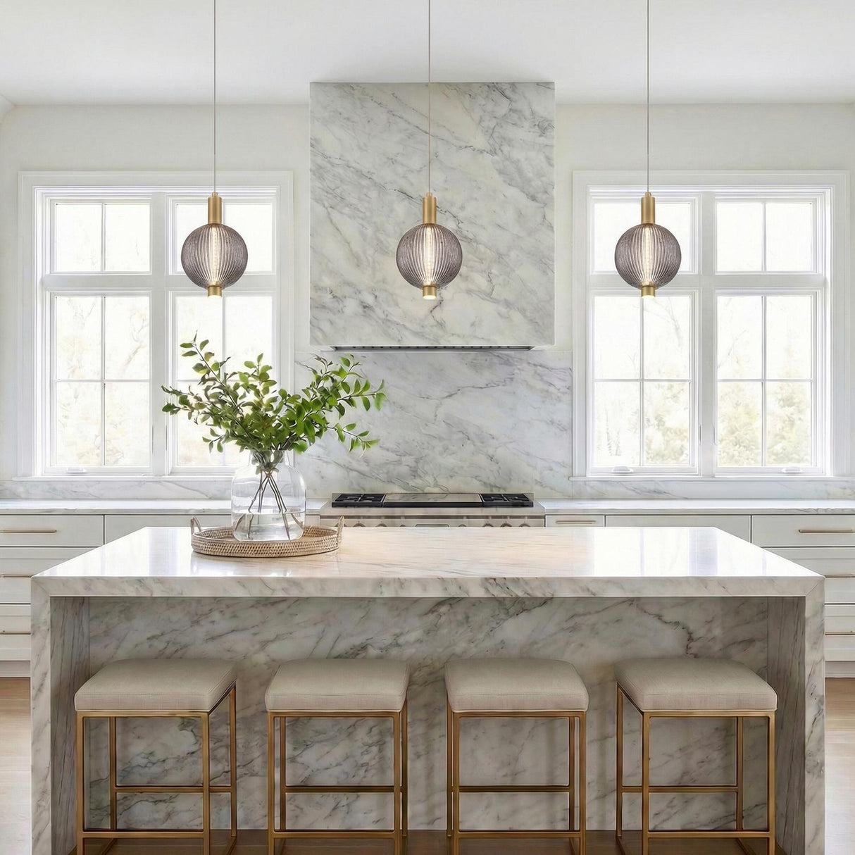 A contemporary kitchen features three Nola LED Pendant 6 inch Brass and Ribbed Smoked Glass lights over a marble waterfall island with four matching brass stools.