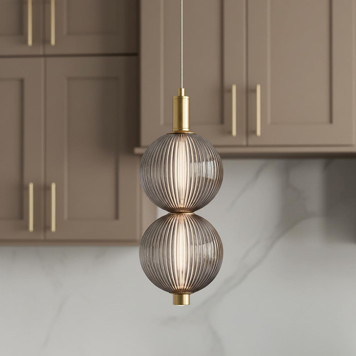 A contemporary kitchen features the Nola LED 2 Light Pendant 6", Brass and Ribbed Smoked Glass suspended over the island against taupe shaker cabinets and marble backsplash.