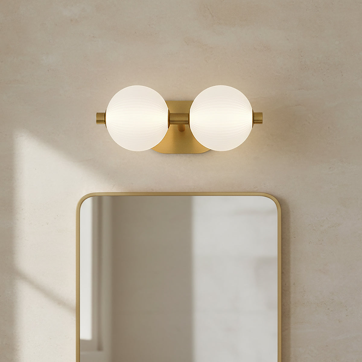 Nola LED 2 Light Vanity 15, Brass and Ribbed Opal Glass - A brass vanity light with two ribbed opal glass globes mounted above a gold-framed mirror against a textured cream wall.