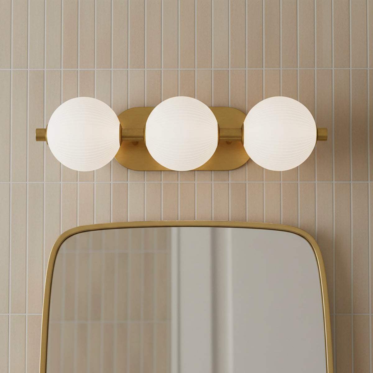 Nola LED 3 Light Vanity 23, Brass and Ribbed Opal Glass - A brass vanity light with three ribbed opal glass globes above a rounded gold mirror, set against a backdrop of vertically stacked beige tiles.