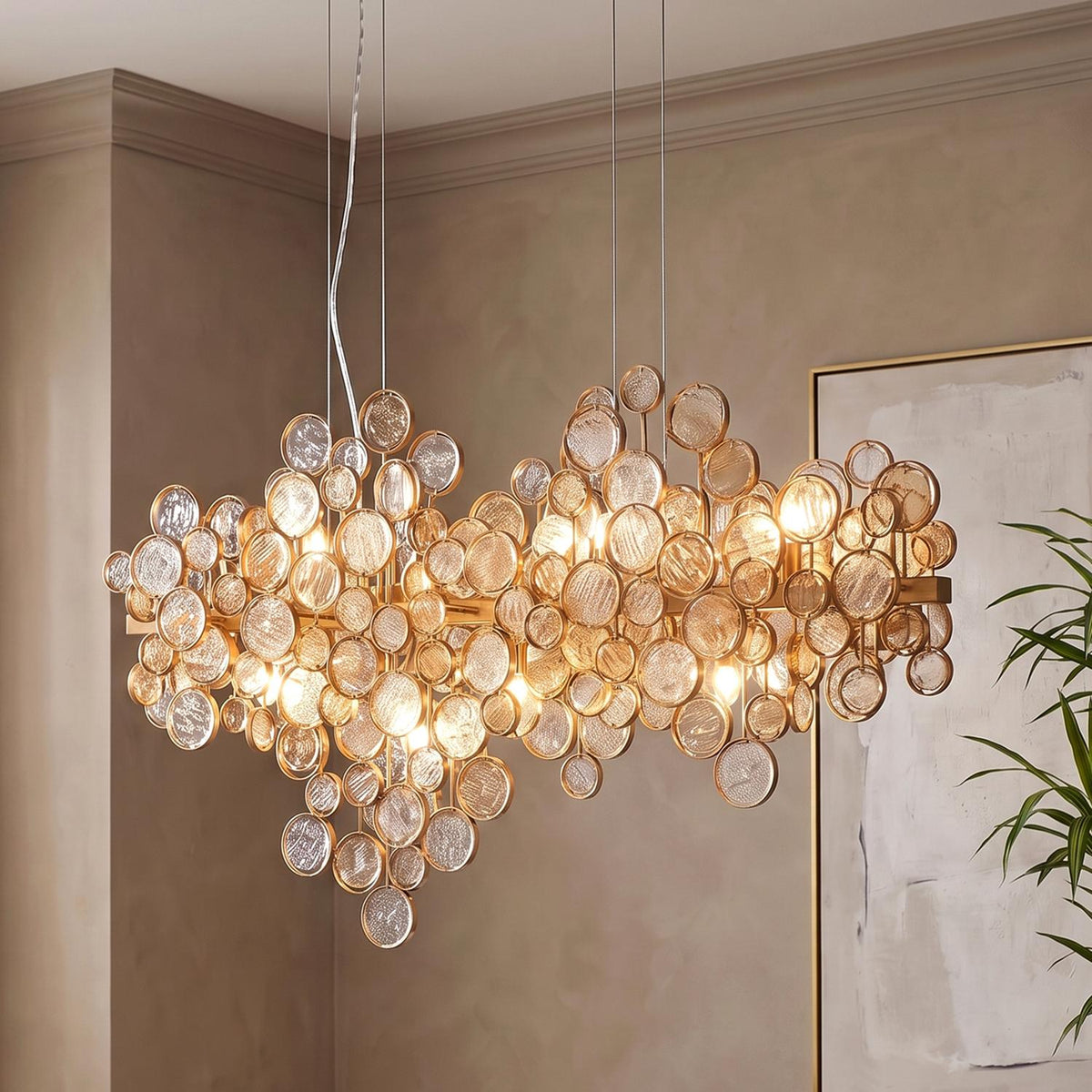 A contemporary dining room featuring the Denali 12 Light Linear Chandelier in antique gold with topaz glass discs suspended near framed wall art and a houseplant.