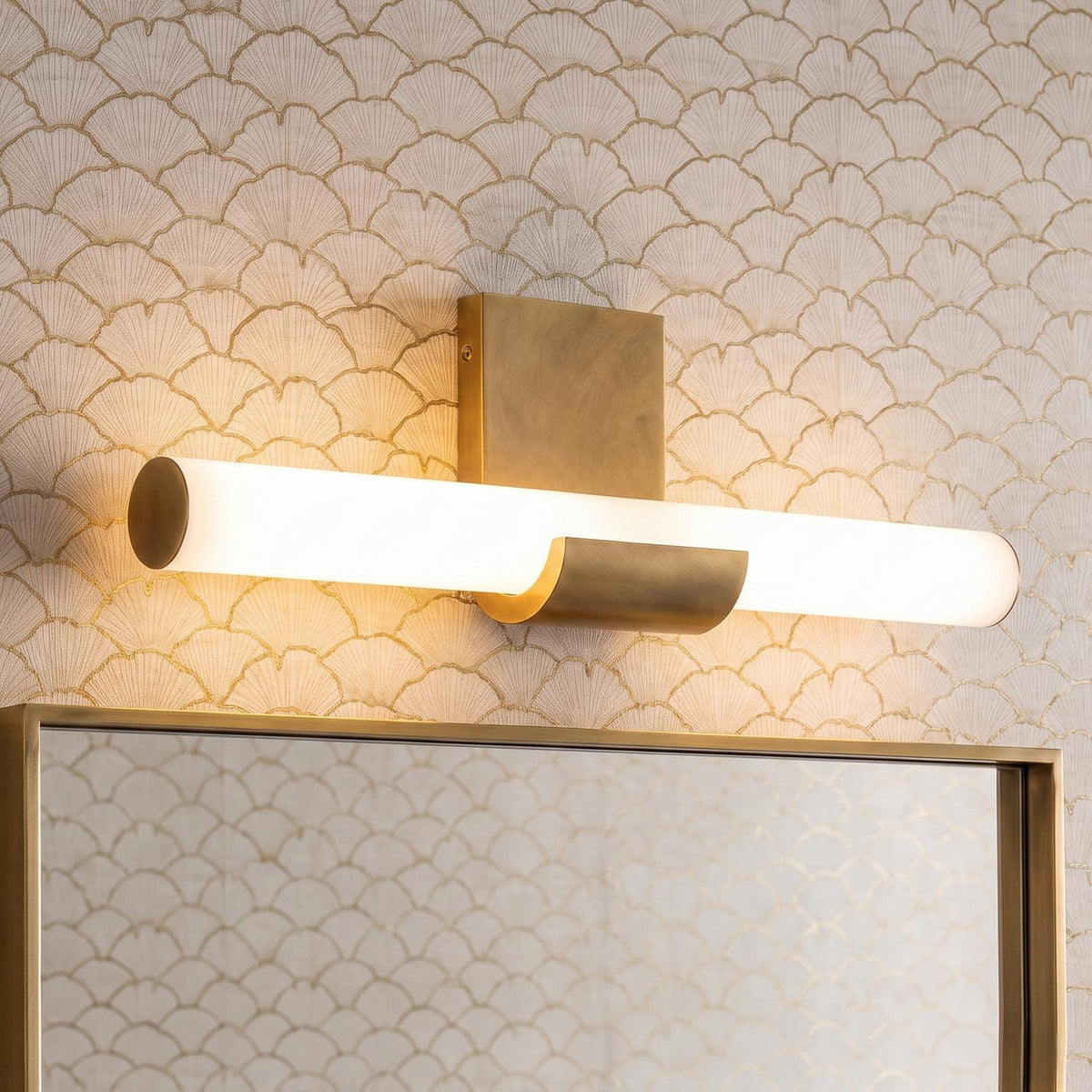 A contemporary bathroom featuring the Nell LED Vanity Light 20", Aged Brass with frosted glass mounted above a brass-framed mirror against metallic fan-patterned wallpaper.