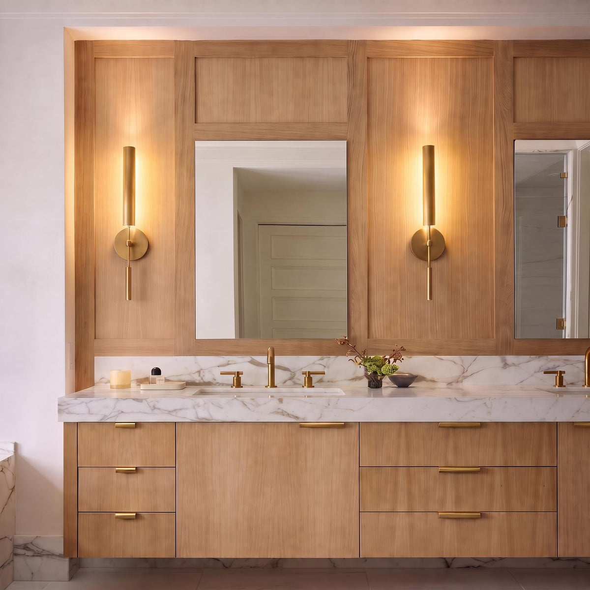 A contemporary bathroom vanity with two Linden LED Wall Sconce 26", Aged Brass fixtures mounted on light wood paneling above a marble countertop.