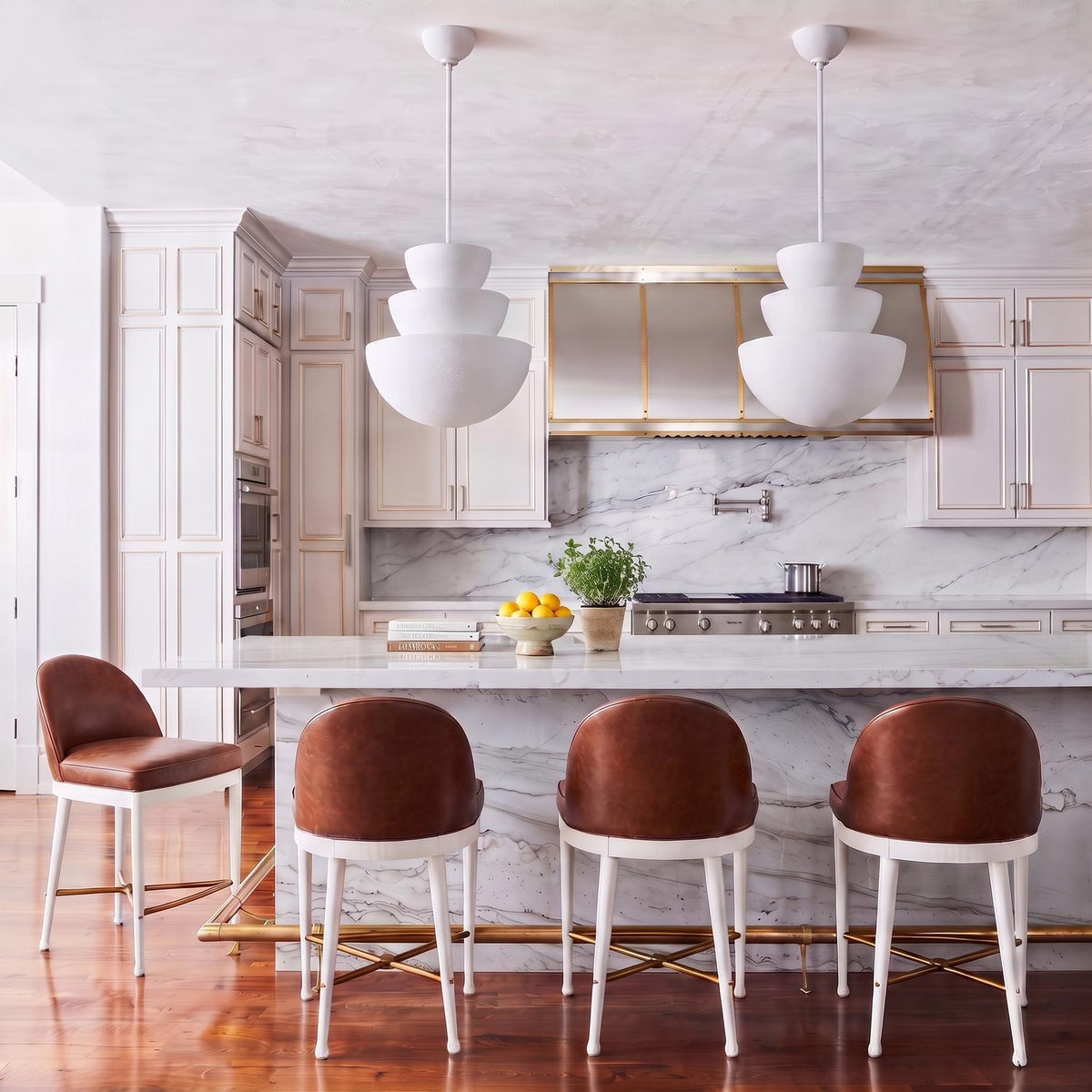 A transitional kitchen with two Verity Pendant 18 inch Textured White pendants over a white marble waterfall island with brown leather bar stools and a brass-trimmed hood.