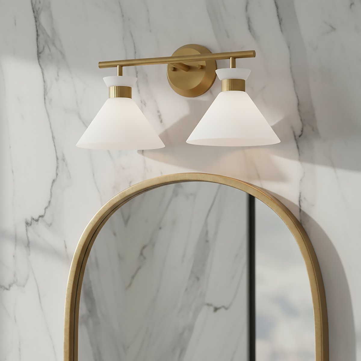Issa 2 Light Vanity, Satin Brass - A modern two-light vanity fixture in satin brass with cone-shaped white glass shades is mounted above an arched mirror with a brass frame.