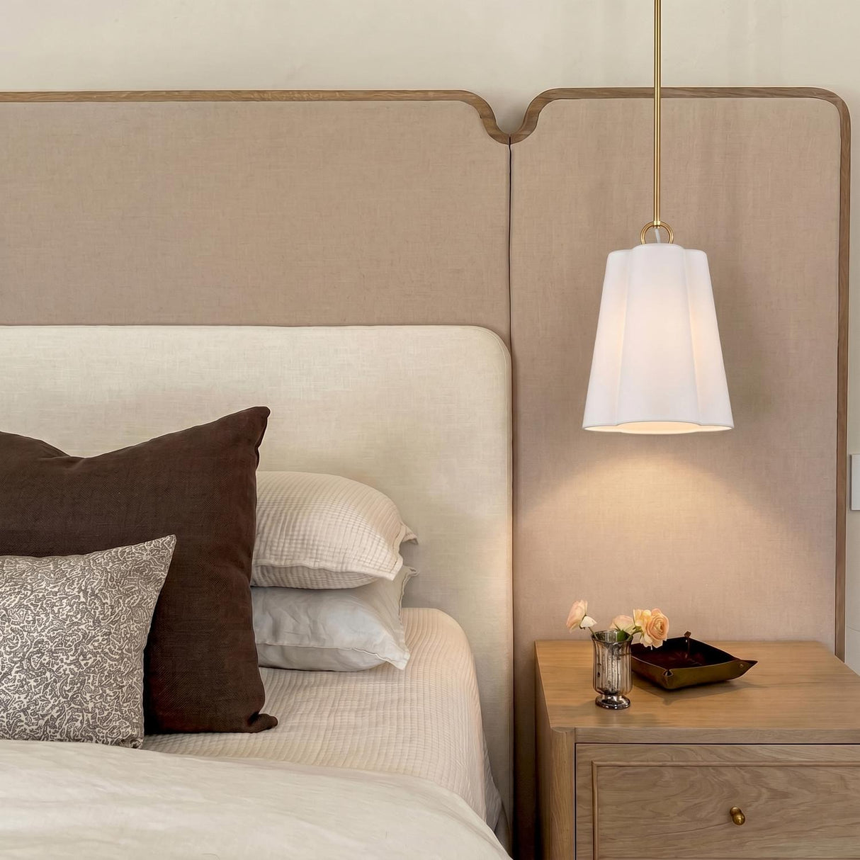 A contemporary bedroom with a white ceramic and brushed brass Odelia Small Pendant hanging over a light wood nightstand beside a large beige fabric headboard.