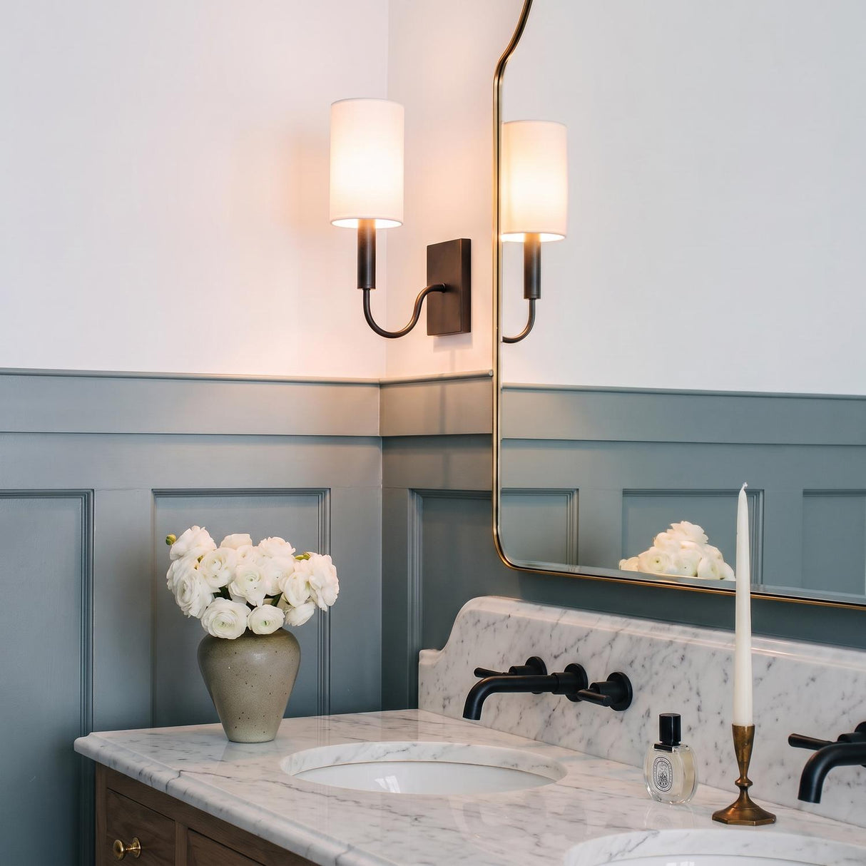A transitional bathroom with an antique iron wall sconce featuring a linen shade beside a gold framed mirror above a white marble countertop and gray wainscoting.