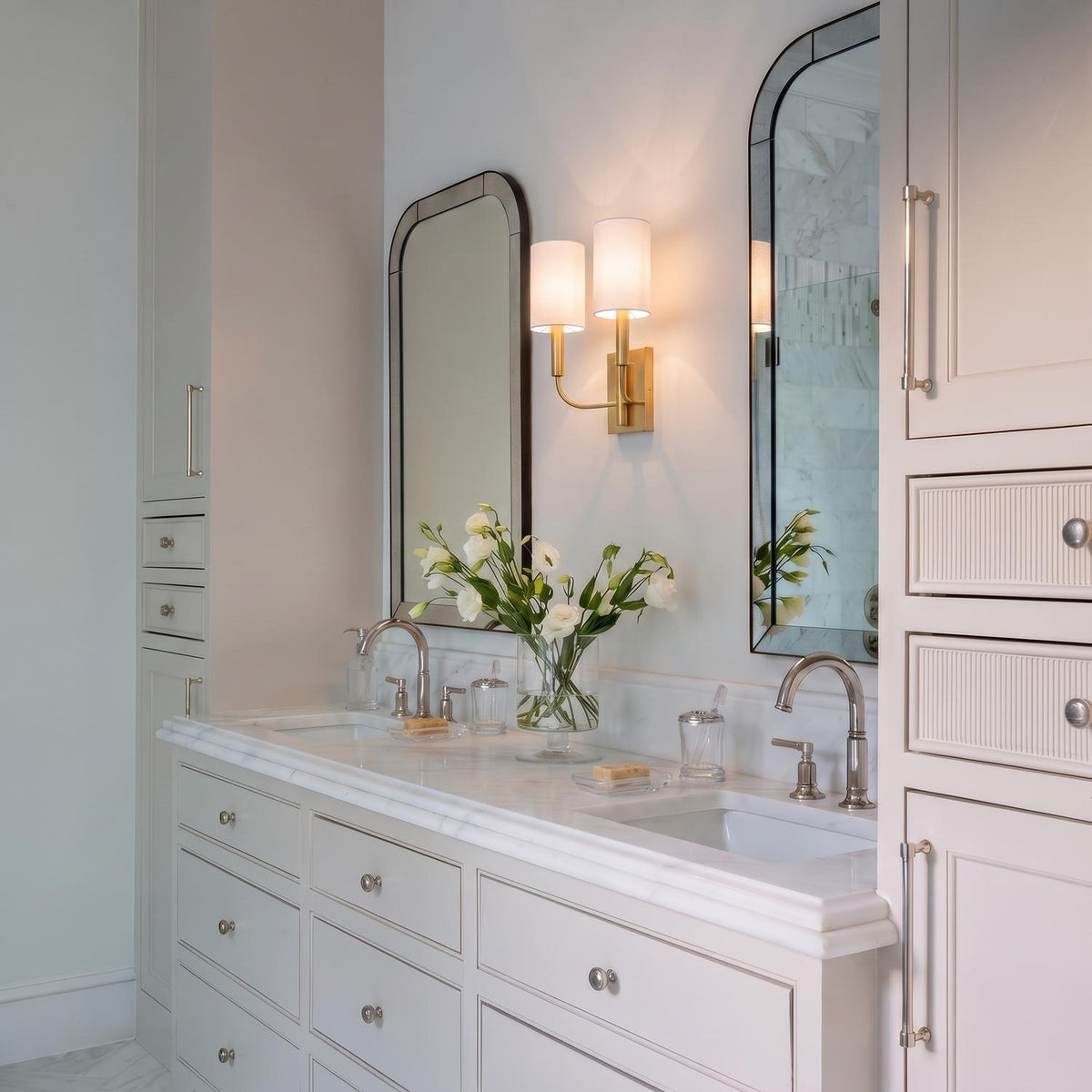 A transitional bathroom featuring a Thayer Double Wall Sconce 14", Modern Brass with linen shades placed between two arched mirrors over a white marble vanity.