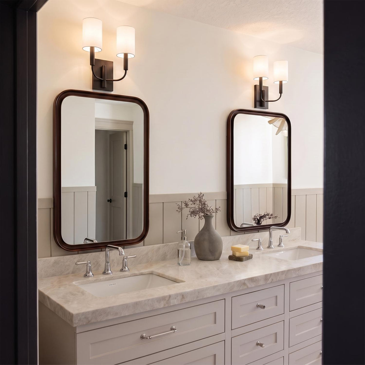 A transitional bathroom with two Thayer Double Wall Sconces in an antique iron finish and linen shades above rounded mirrors, a marble vanity, and wainscoting.