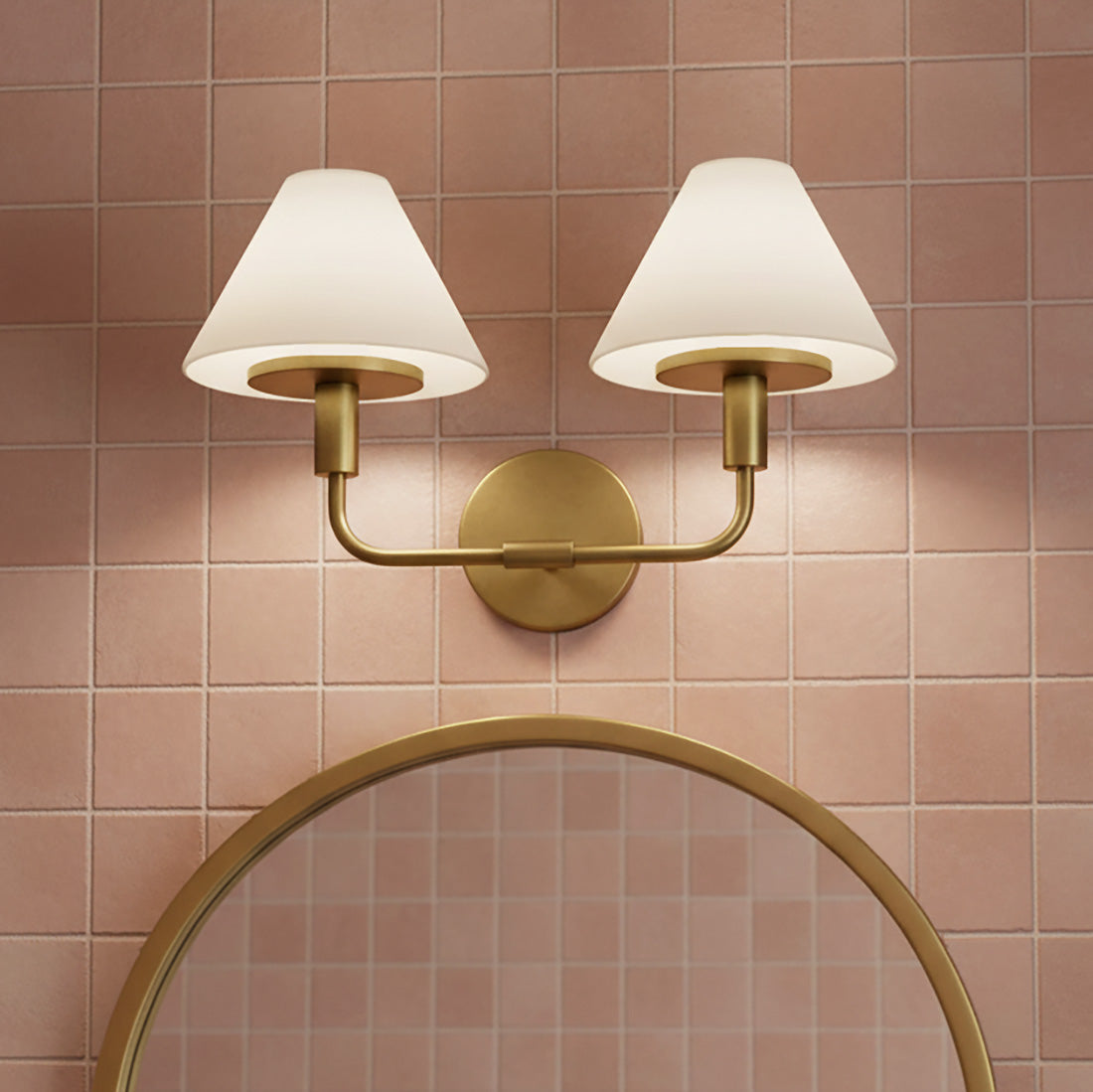 Dean 2 Light Vanity - A brass and opal glass vanity light above a round gold mirror against a pink tiled wall.