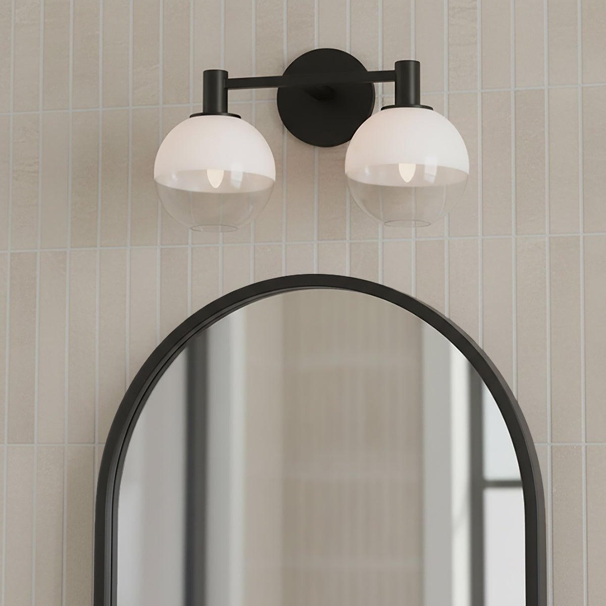 Merrick 2 Light Vanity, Aged Iron - A black two-light vanity fixture with globe shades is mounted above an arched mirror on a wall with vertical cream tiles.