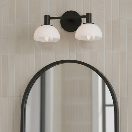 Merrick 2 Light Vanity, Aged Iron - A black two-light vanity fixture with globe shades is mounted above an arched mirror on a wall with vertical cream tiles.