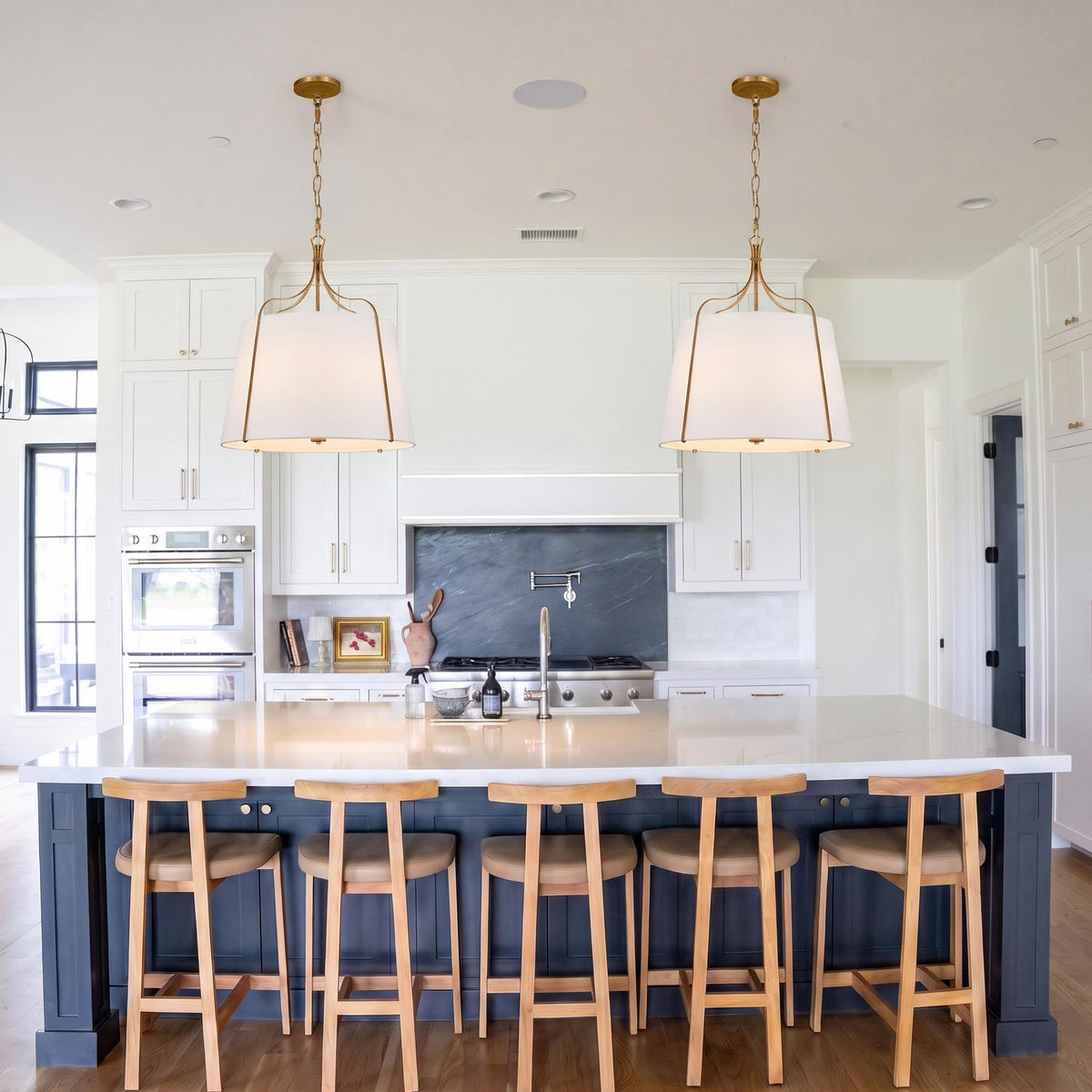 A transitional kitchen featuring two Isleton Large Pendants in aged gilded brass with linen shades over a white quartz island and dark navy blue cabinetry.