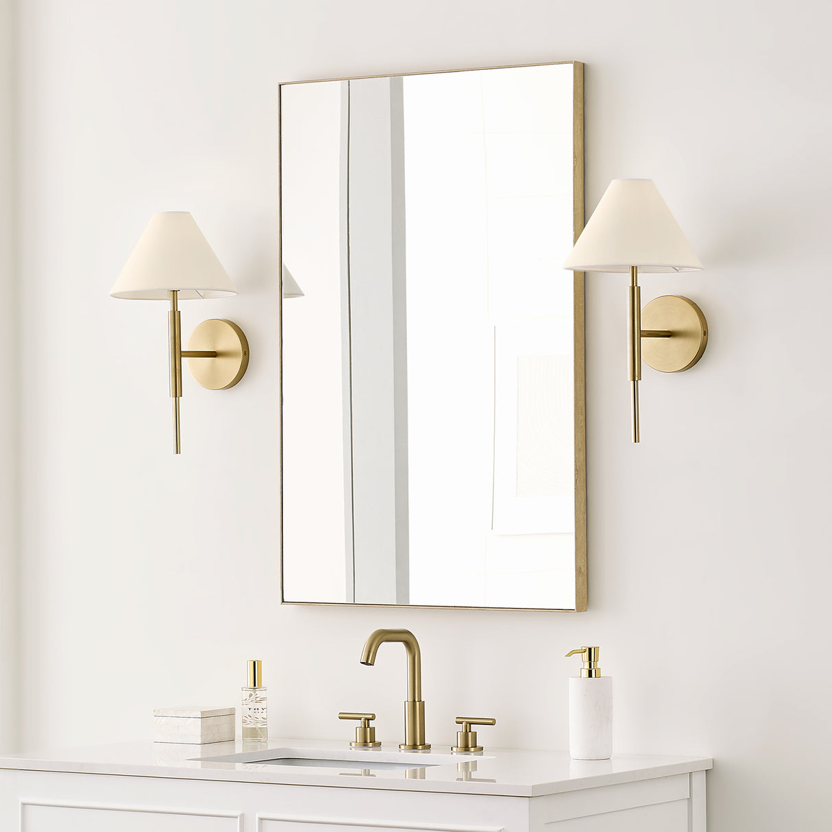 Two 17" tall Hazelton Wall Sconces in Satin Brass with white fabric shades flanking a rectangular gold framed vanity mirror in a bright bathroom