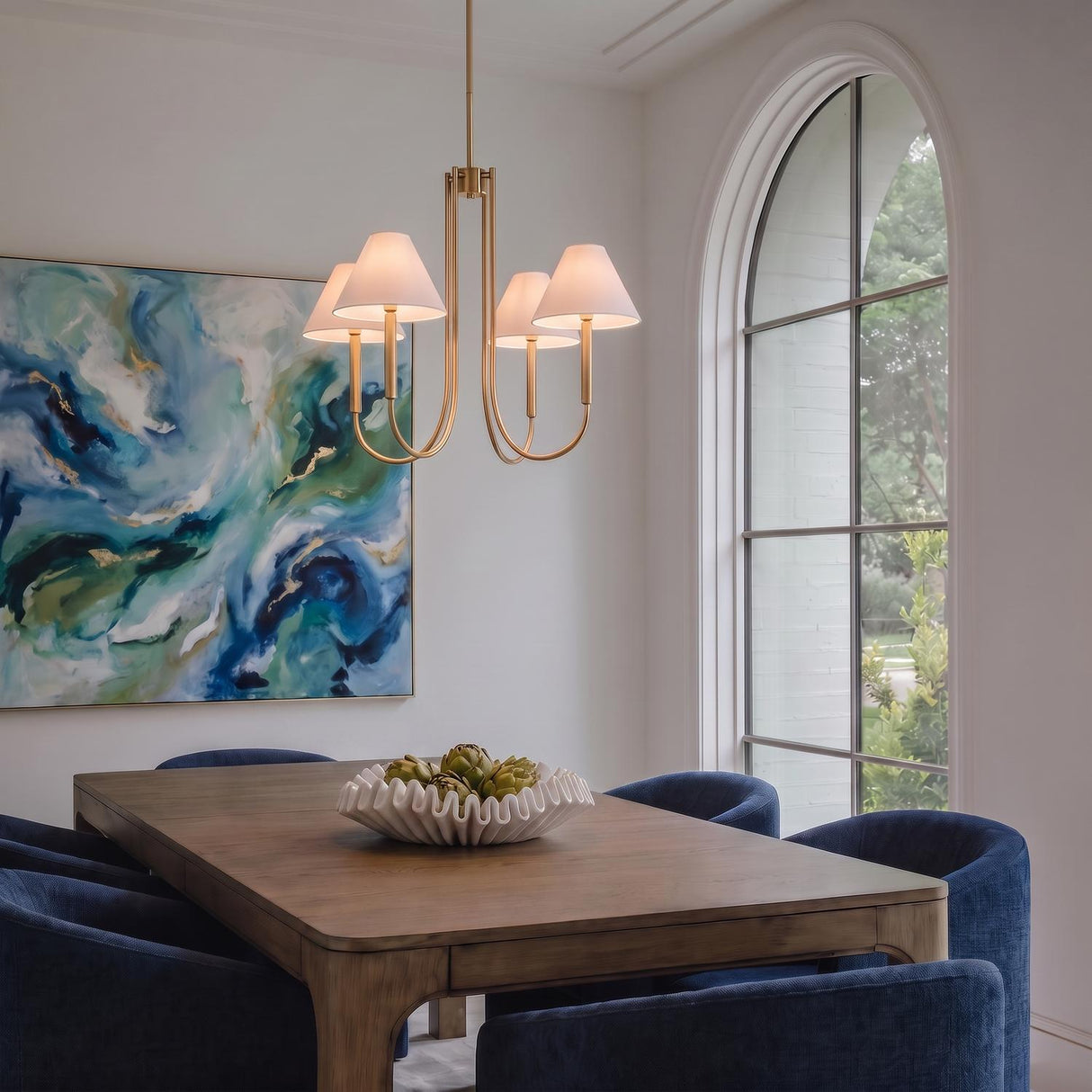 A transitional dining room featuring the Hazelton 4 Light Chandelier 30 inch in satin brass over a wood table with blue velvet chairs and abstract art.