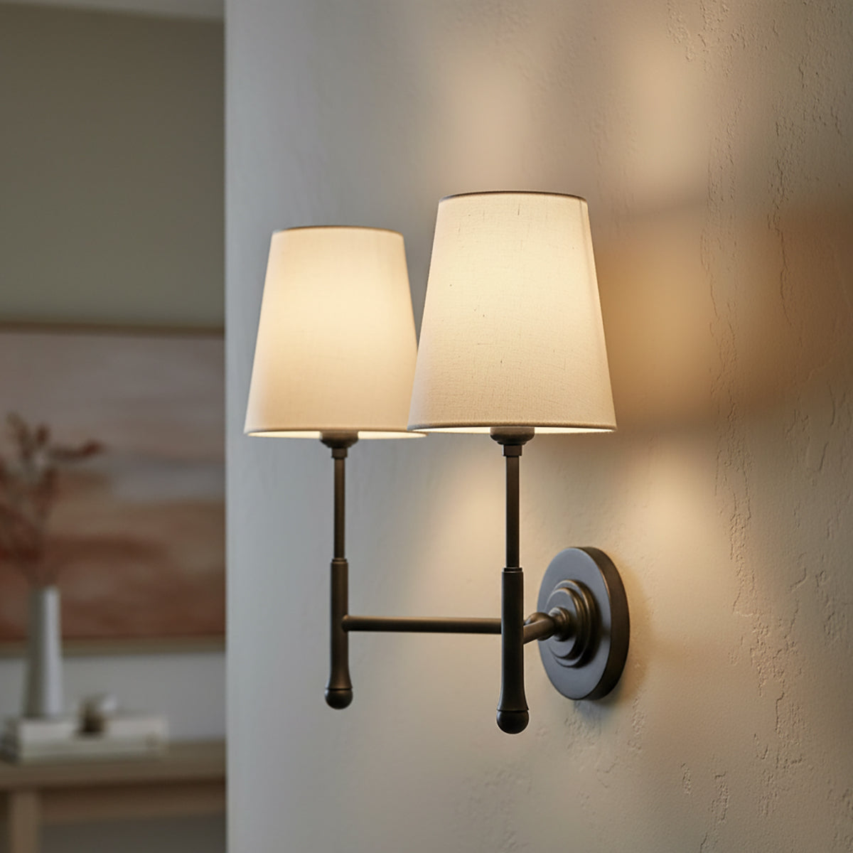 Emerson Double Wall Sconce 14 - A double wall sconce with aged iron finish and cream linen shades is mounted on a textured wall, casting warm light.