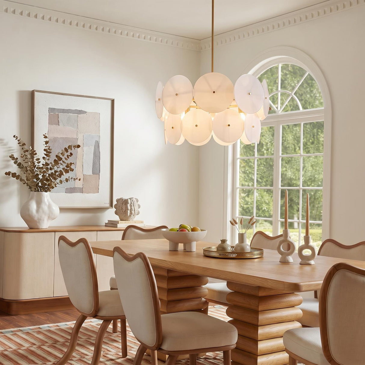 A contemporary dining room featuring the Ophelia Medium Chandelier with modern brass and layered glass discs hanging over a wood pedestal dining table and cream chairs.