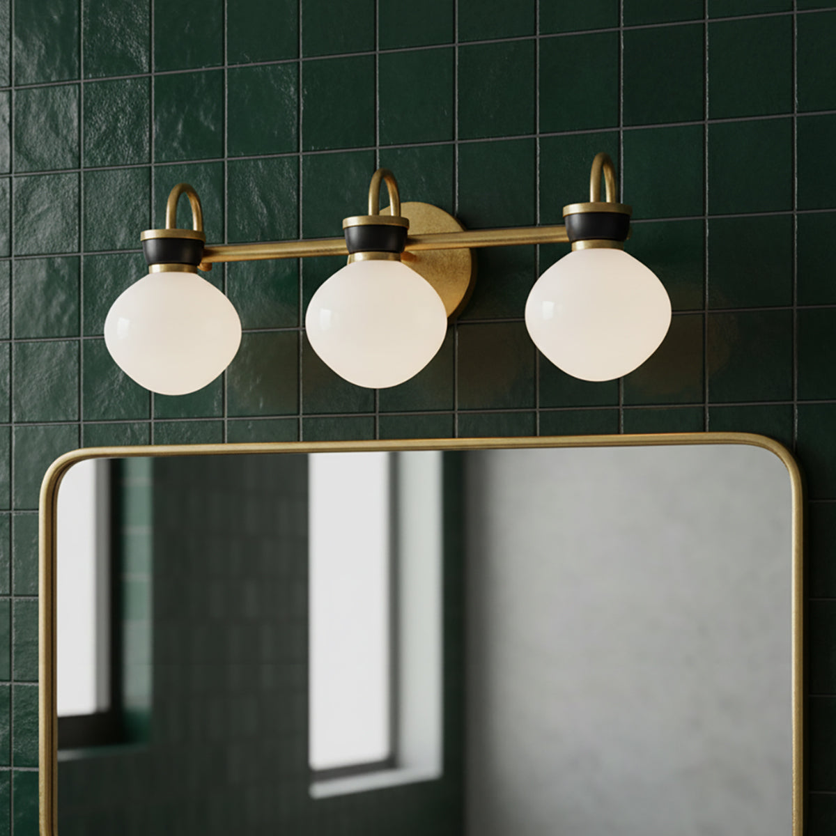 Piper 3 Light Vanity, Satin Brass and Black - A brass vanity light with three opal globes hangs above a gold-framed mirror against a dark green tiled wall.