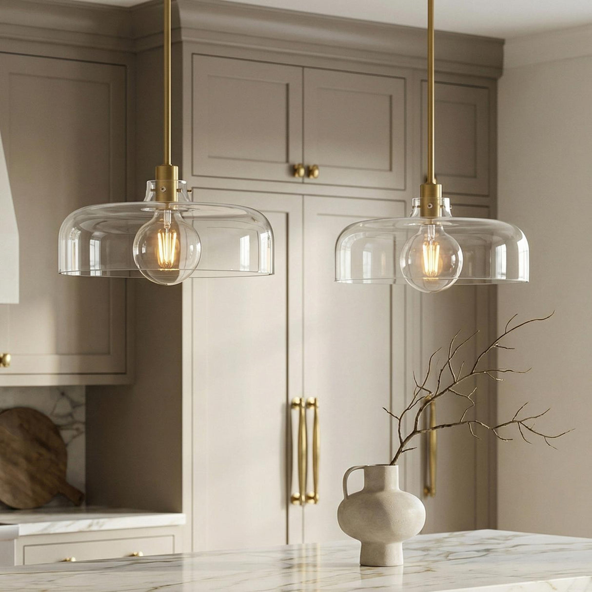 A transitional kitchen with two Alder Pendant 15 inch fixtures in satin brass and clear glass hanging over a white marble island with taupe cabinets.