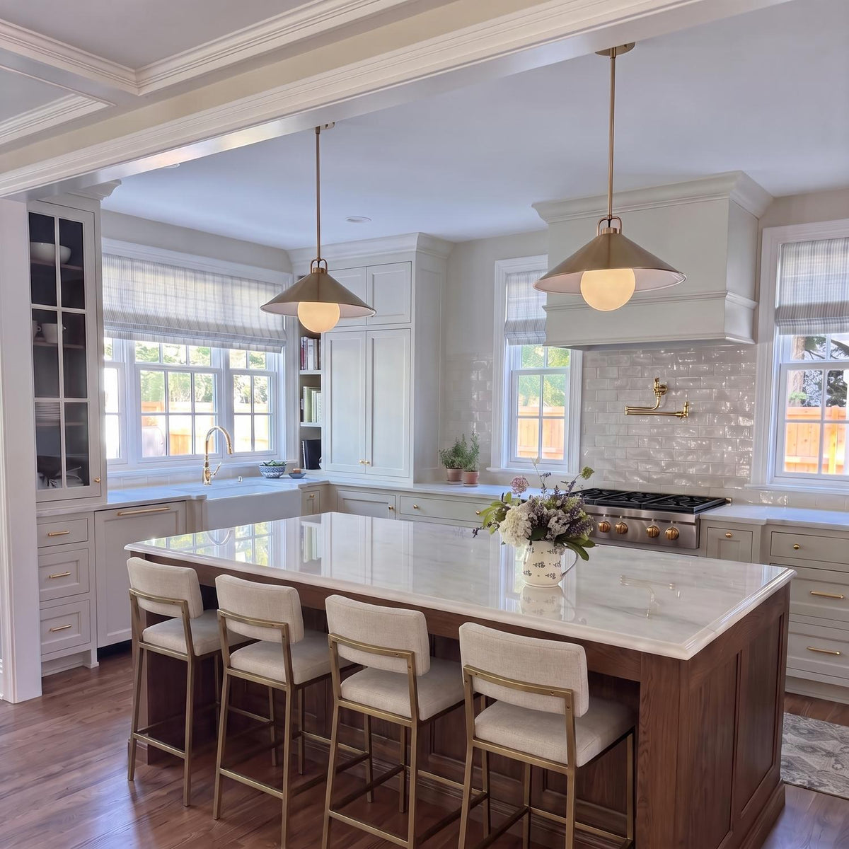A transitional kitchen features two Nyra Pendant 16", Satin Brass lights with white glass globes suspended over a marble island surrounded by off-white cabinets.