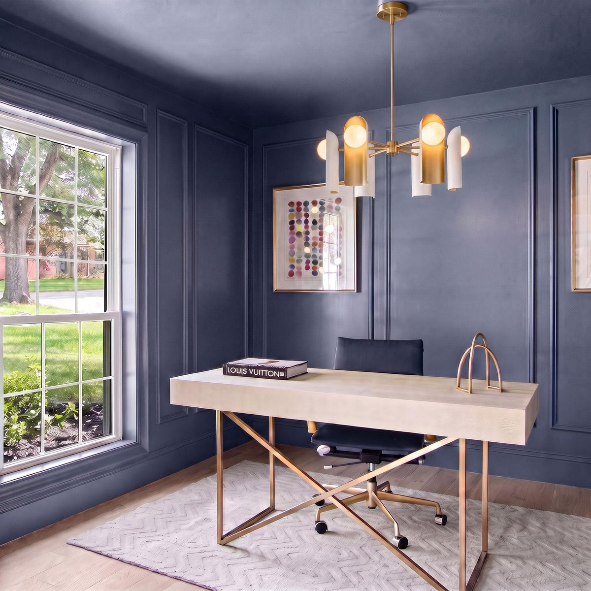 A contemporary office with dark blue paneled walls displays the Kassidy six-light chandelier in satin brass and white above a beige desk and patterned rug.