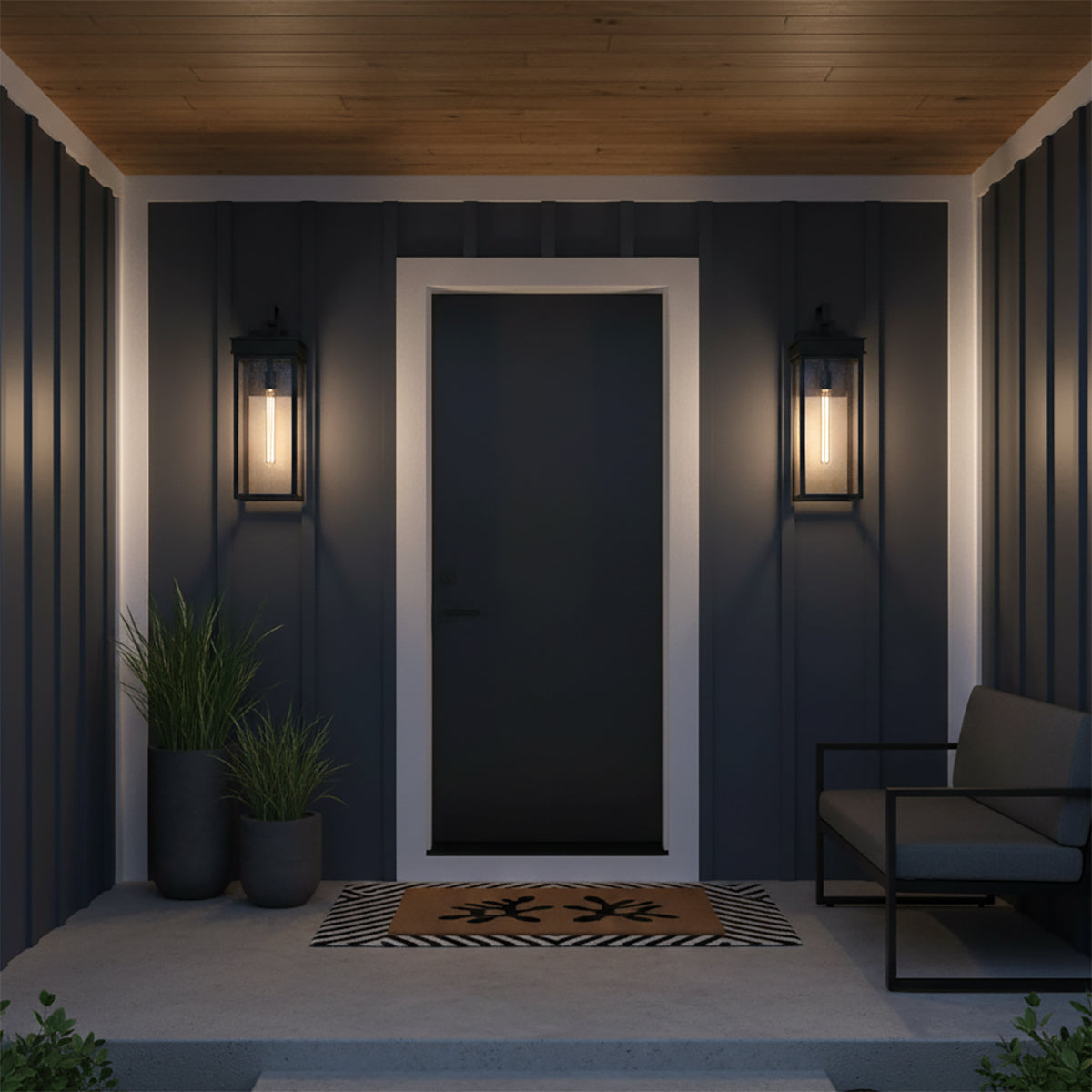 Monterey Outdoor Wall Light, Large 25" Black - Two black lanterns flanking a dark gray door on a covered porch with wooden ceiling, a bench, plants, and a doormat.