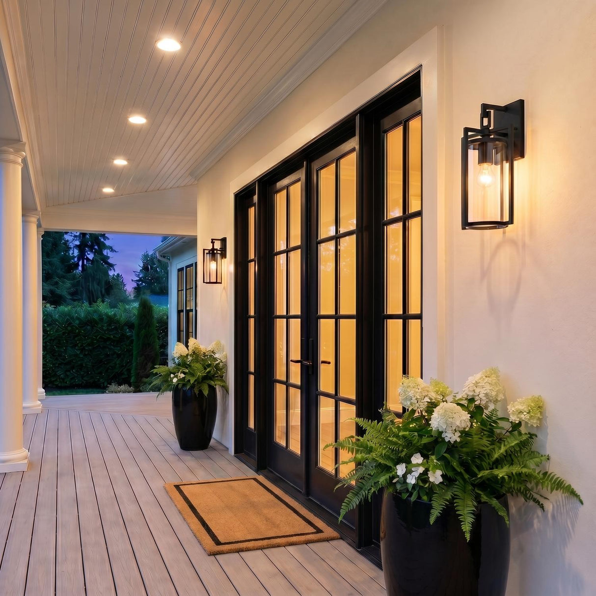 A transitional porch featuring the Peyton Outdoor Wall Light, Small 15" Textured Black flanking black glass doors on white stucco at dusk with potted hydrangeas.