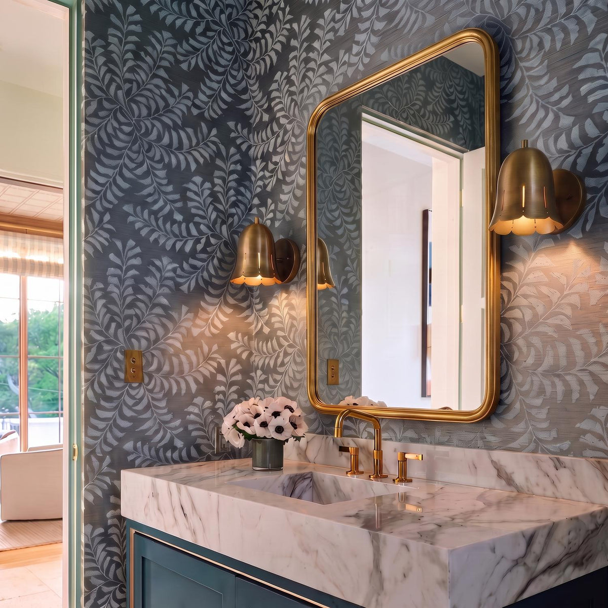 A transitional bathroom features two Sandune Wall Sconce 7", Patina Brass fixtures flanking a curved brass mirror above a white marble vanity set against blue patterned wallpaper.