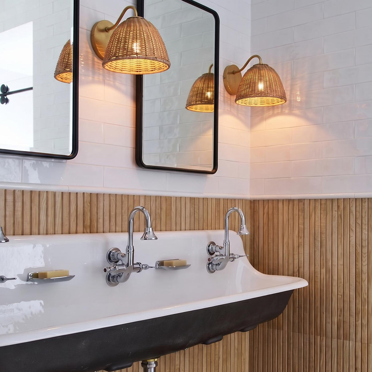 A coastal bathroom with two Karina Wall Sconce 8" fixtures in woven wicker and aged brass flanking mirrors above a white trough sink with chrome faucets.