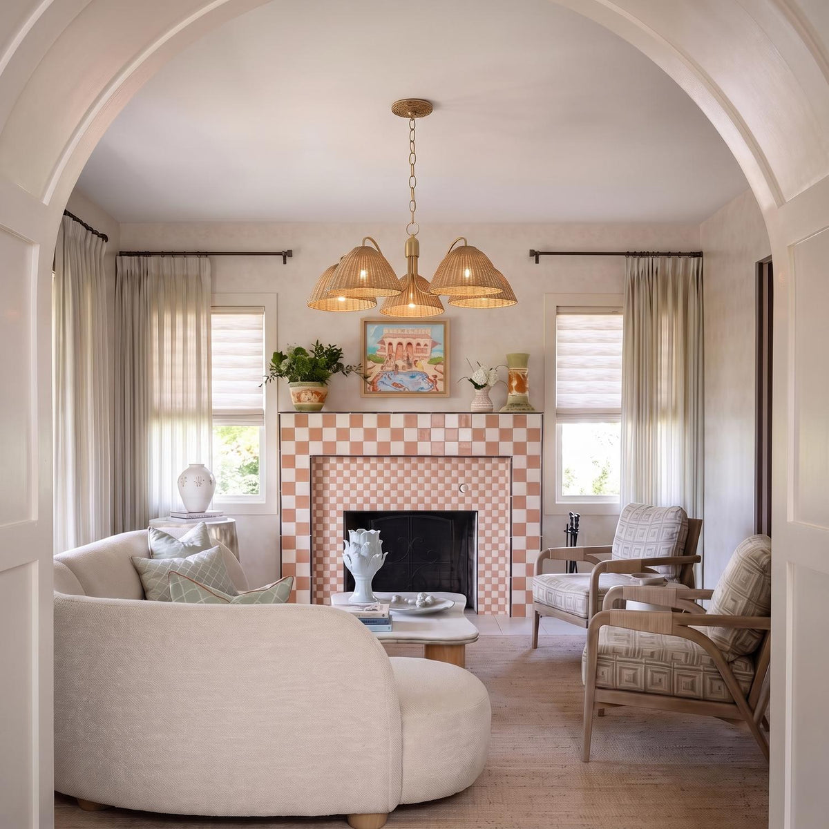 A transitional living room features a Karina 5 Light Chandelier in woven wicker and aged brass suspended above a pink checkered tile fireplace and curved cream sofa.