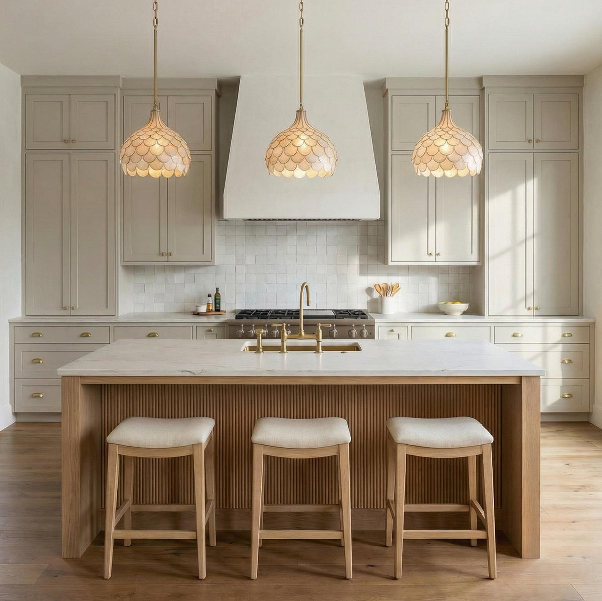 A transitional kitchen with three Valentina Medium Pendant 14", Soft Brass and Capiz Shell fixtures over a fluted wood island with white marble and taupe cabinets.