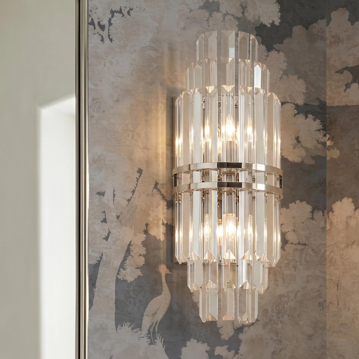 A traditional foyer with a Maxwell Large Wall Sconce 20" in crystal and polished nickel mounted on scenic tree-patterned mural wallpaper beside a mirror.