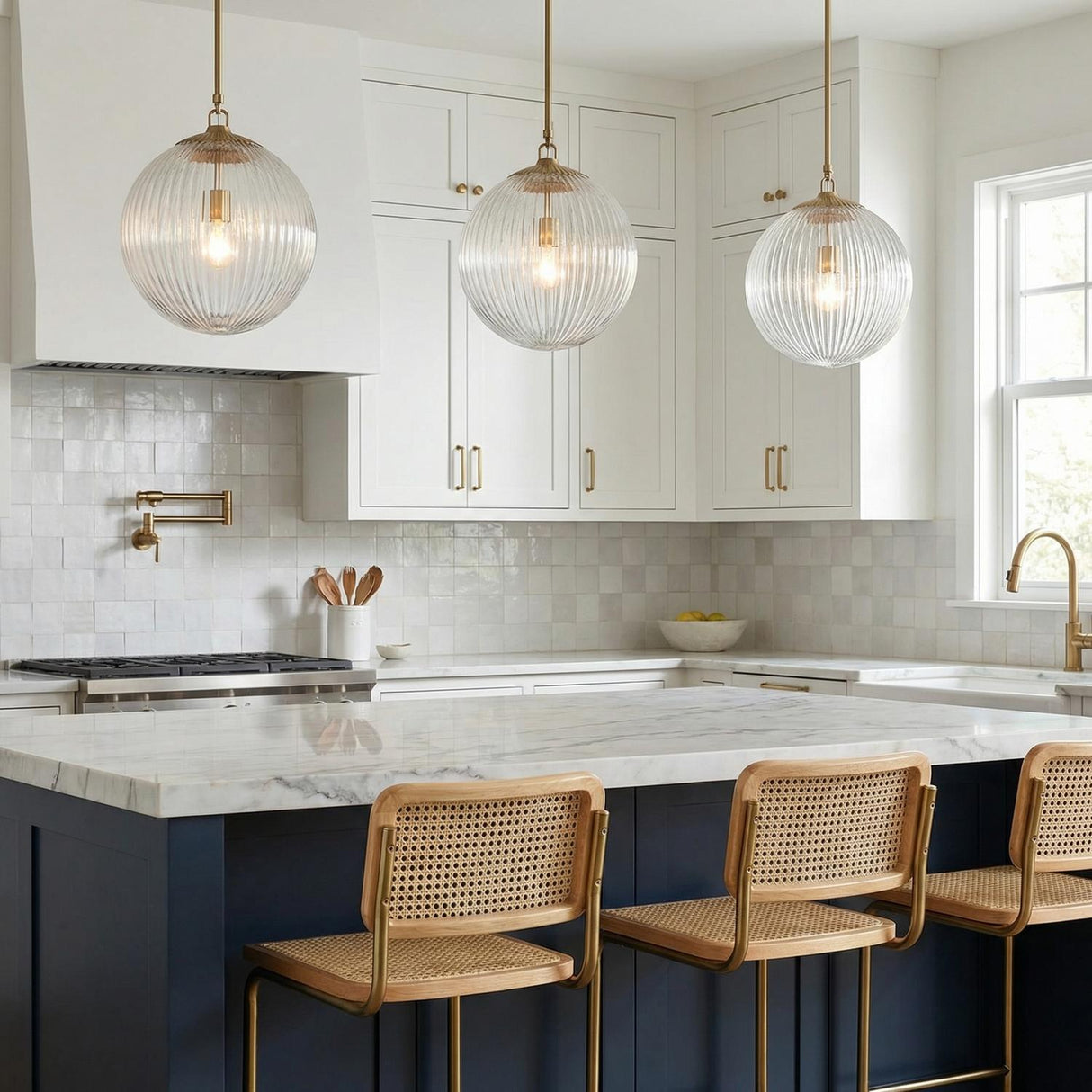 A transitional kitchen features three Victoria Pendant 14", Aged Brass and Clear Ribbed Glass fixtures over a marble island with navy cabinets and rattan stools.