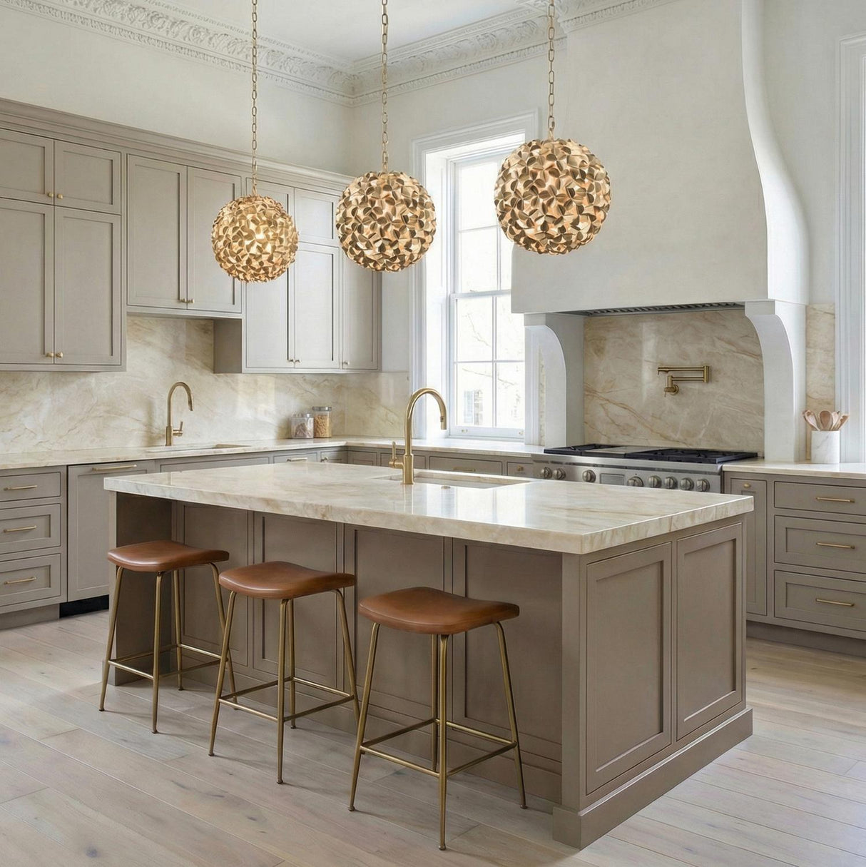 A transitional kitchen features three Florence Pendant 13", Soft Brass globe fixtures over a marble island surrounded by taupe cabinetry and a matching stone backsplash with brass faucets.