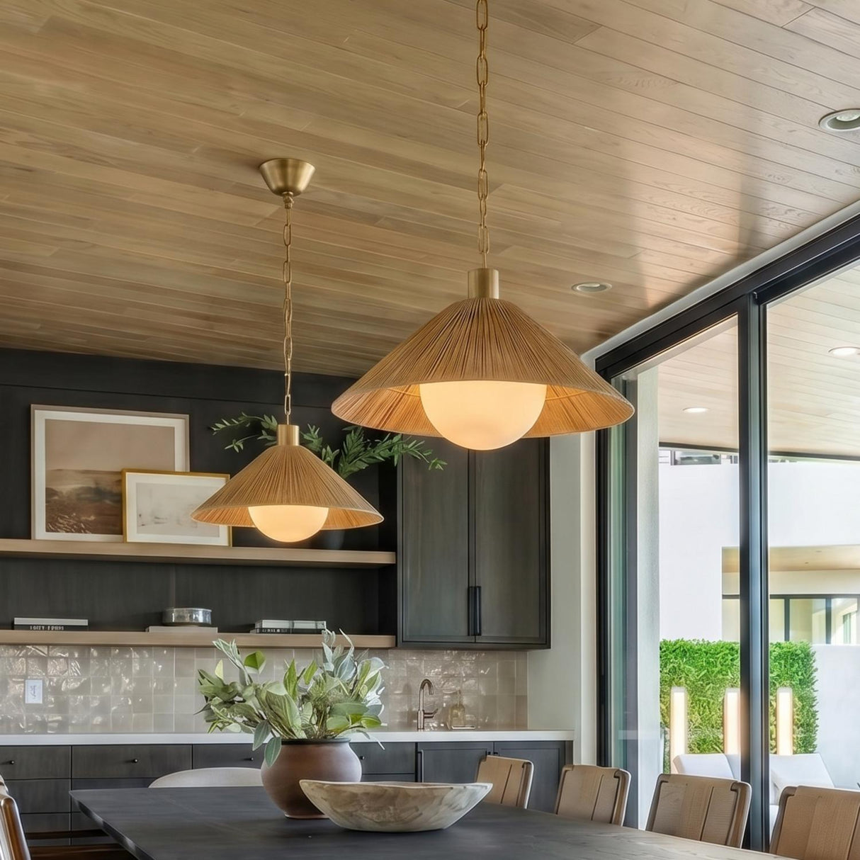 A modern dining room with two Fawn Medium Pendants in patina brass and rattan over a dark wood dining table against dark cabinets and a wood-planked ceiling.