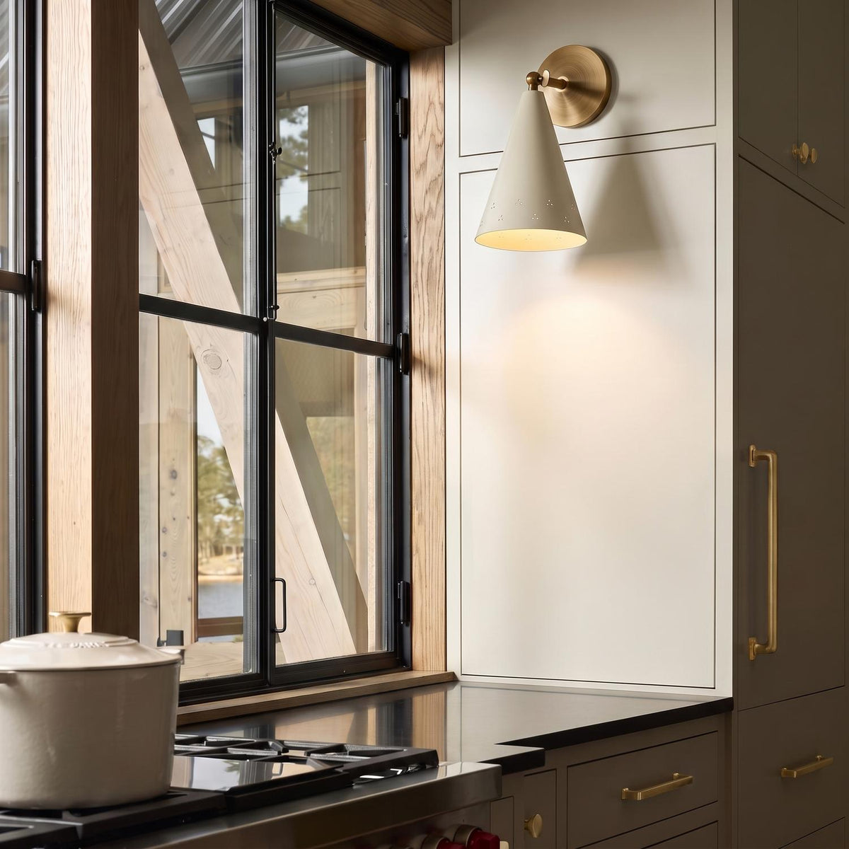 A modern farmhouse kitchen featuring a soft sand and patina brass cone wall sconce mounted on cream cabinetry beside a black window and stainless steel range.