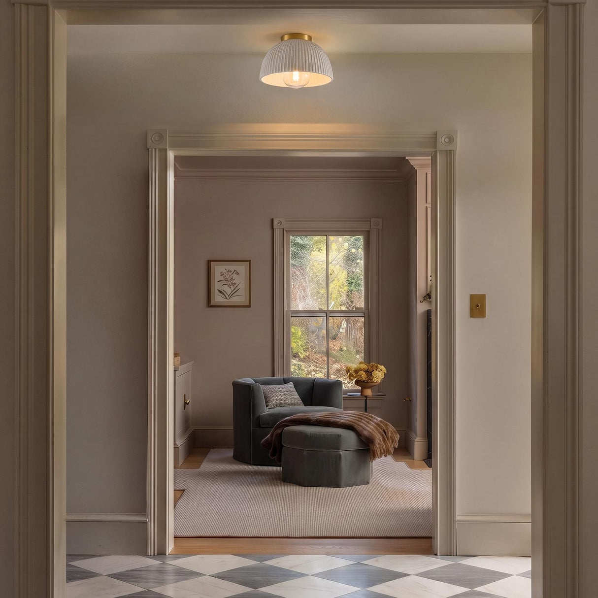 A traditional foyer with the Pippa Flush Mount 12", Aged Brass and Rustic White along the ceiling over checkered marble tile and a grey velvet armchair.