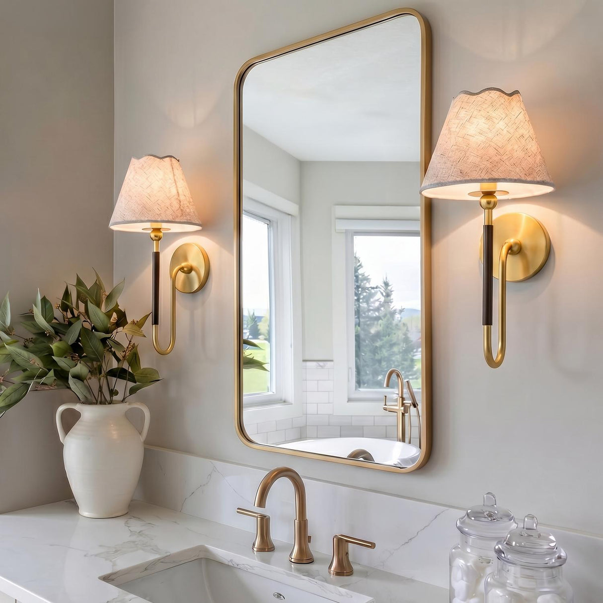 A transitional bathroom with two Sara Wall Sconce 17", Aged Brass and Bronze wall sconces with linen shades flanking a gold framed mirror above a marble vanity.