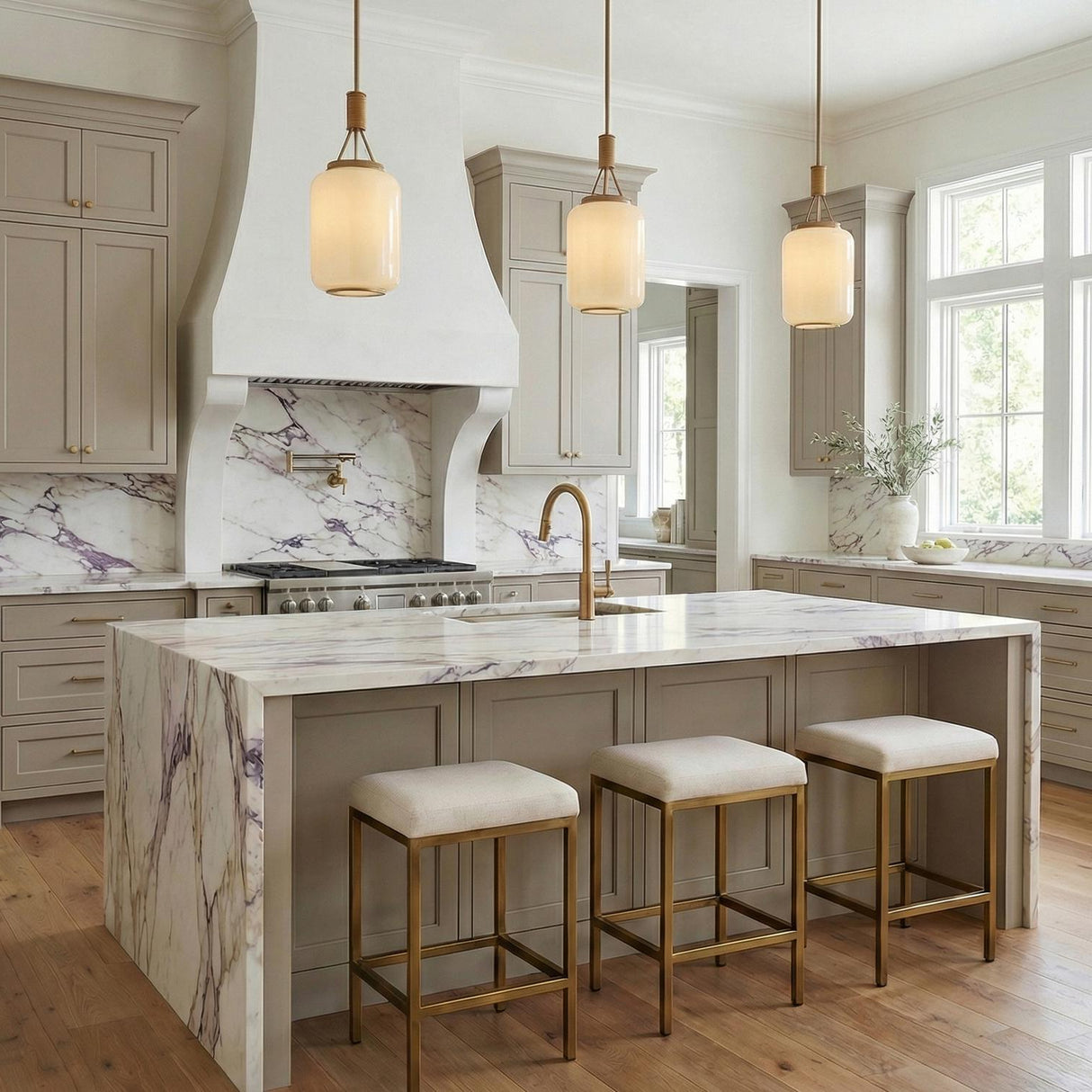 A transitional kitchen features three Yuma Pendant 10-inch, Patina Brass and Rattan with Cream Milk Glass pendants over a purple veined marble waterfall island and taupe cabinets.
