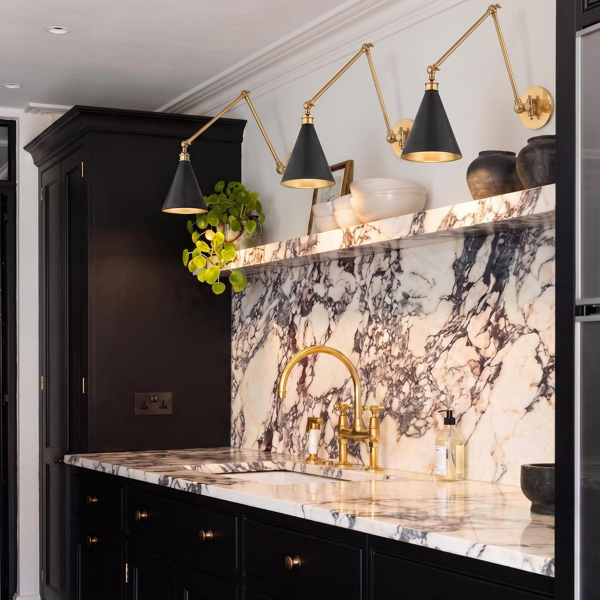 A contemporary kitchen featuring three Ambrose Articulating Wall Sconces in Urban Bronze and Aged Brass over a dramatic marble backsplash with integrated shelf and black cabinetry.