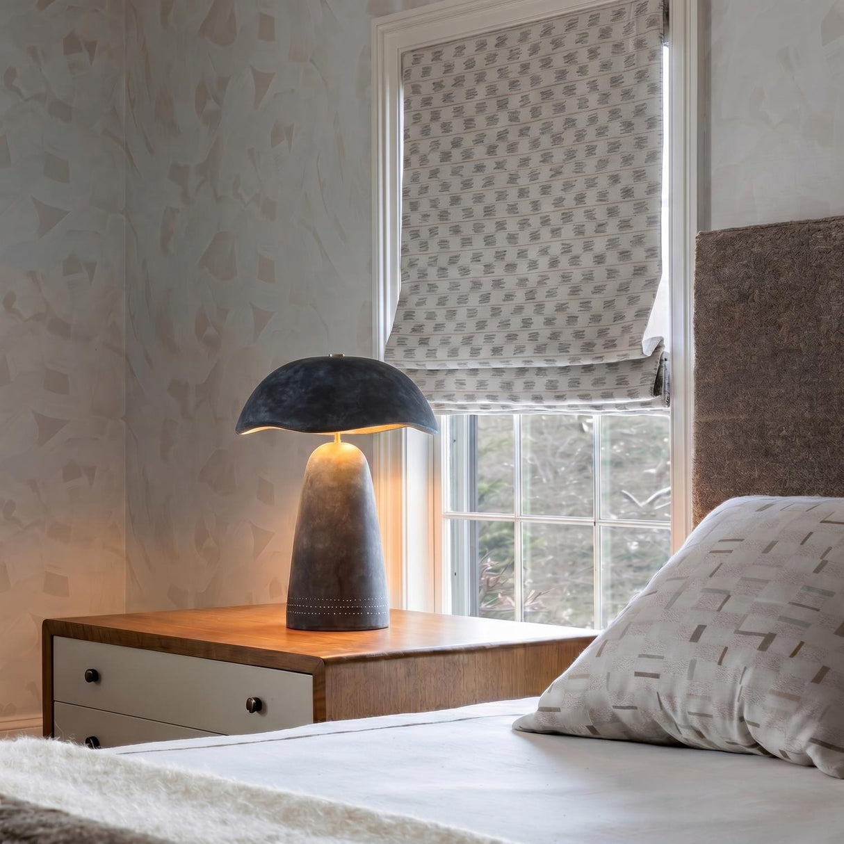 A contemporary bedroom with the Janna Table Lamp 21 in Artisan Slate on a wood nightstand featuring patterned wallpaper and a neutral Roman shade.
