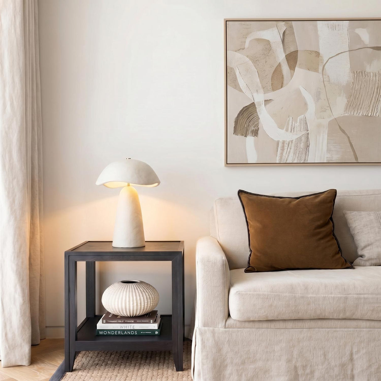 A contemporary living room featuring the Janna Table Lamp 21", Artisan Ivory on a dark wood side table next to a beige sofa and abstract wall art.