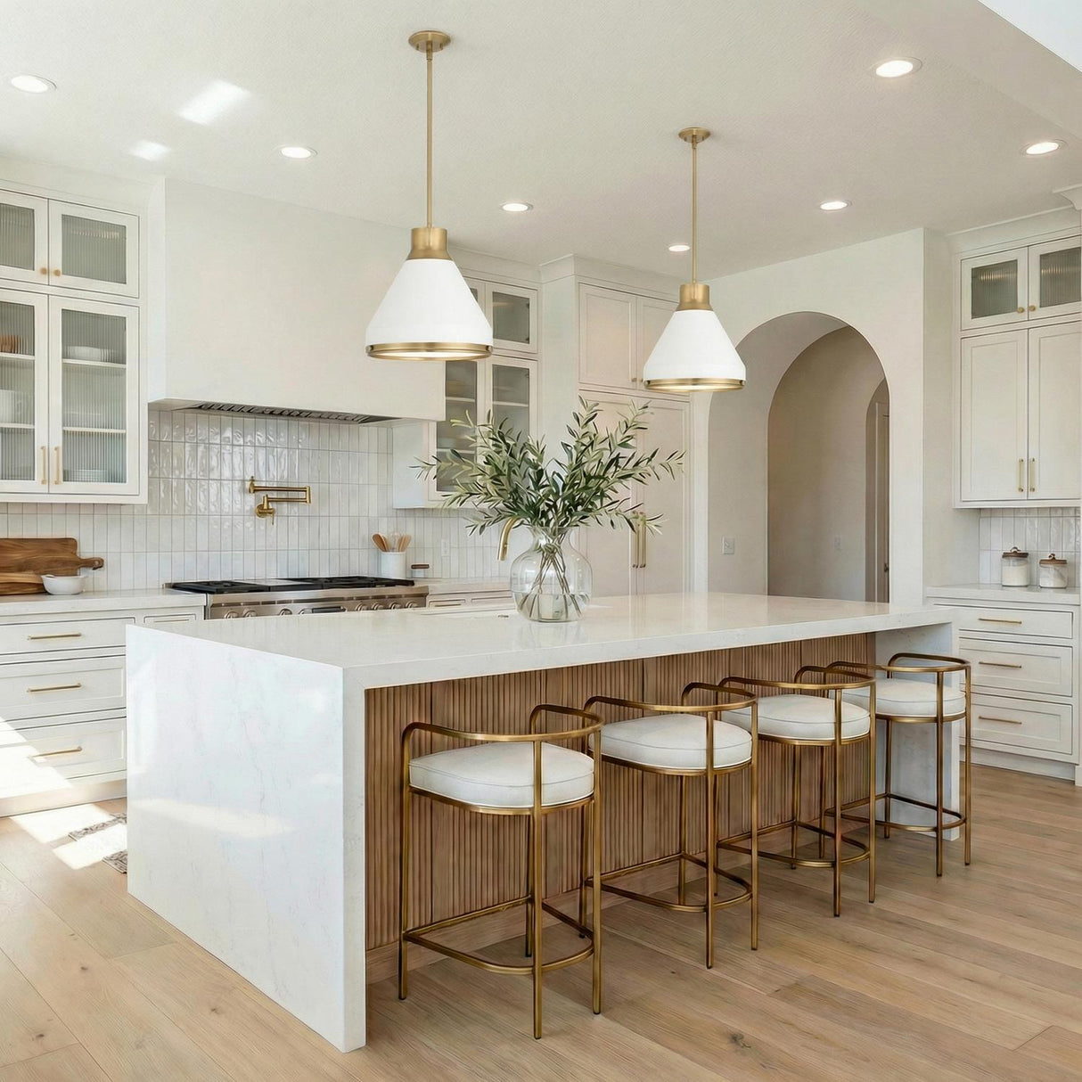 A contemporary kitchen features the Drew Medium Pendant 19 in matte white and aged brass over a white quartz waterfall island with wood fluting and brass barstools.