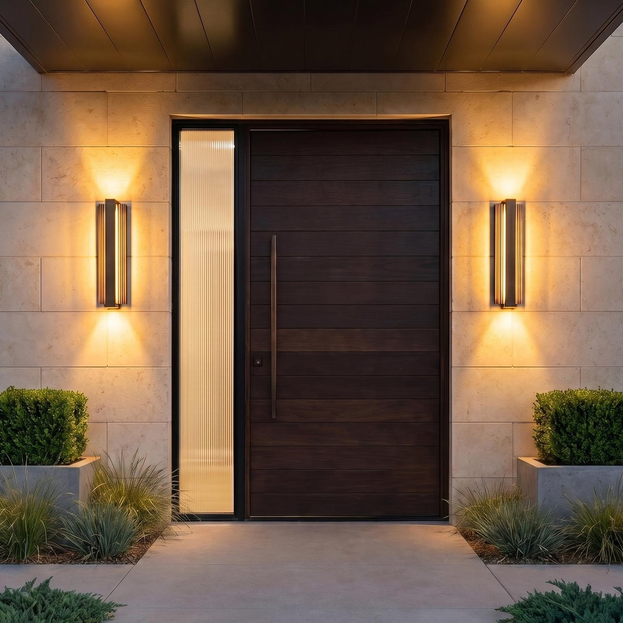 A modern home entry with two Zenith Outdoor LED Wall Light, Large 24", Bronze fixtures flanking a dark wood door on stone walls at night with shrubs.