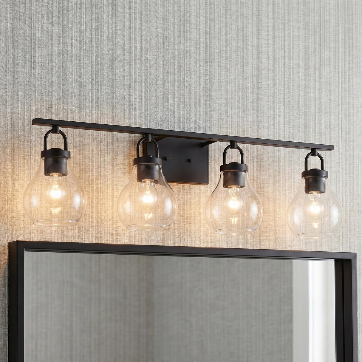 A transitional bathroom features a Naya 4 Light Vanity in matte black and clear glass installed above a black framed mirror against a textured gray wallpaper background.