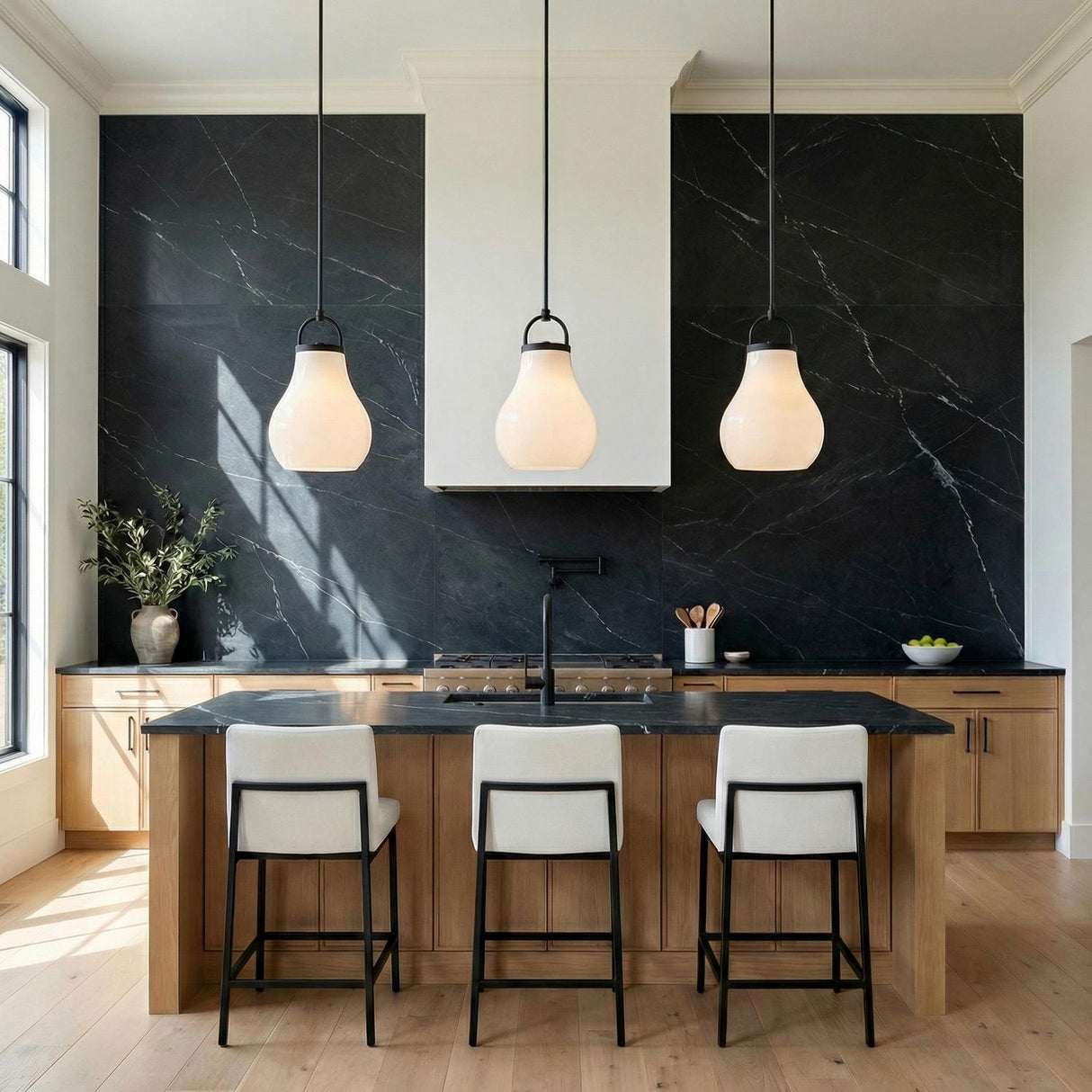 A contemporary kitchen features three Naya Medium Pendant 12", Matte Black and Opal Glass fixtures suspended over a black marble island with wood cabinetry and backsplash.