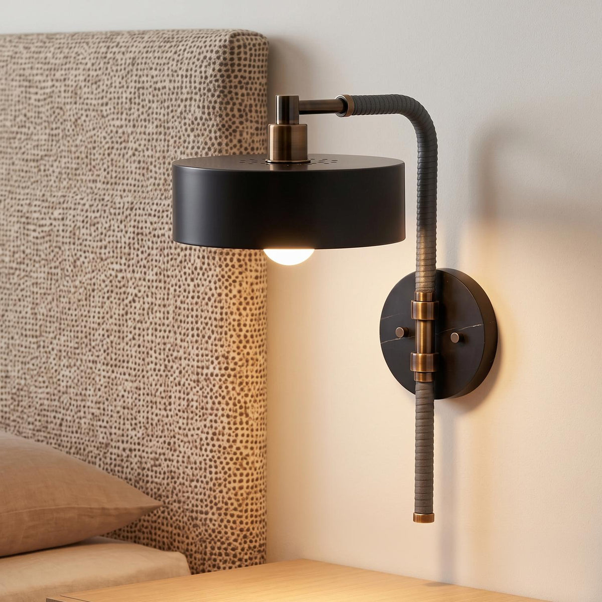 A modern bedroom features an Amadeo Wall Sconce 18", Black and Bronze mounted on a white wall beside a patterned upholstered headboard and light wood nightstand.