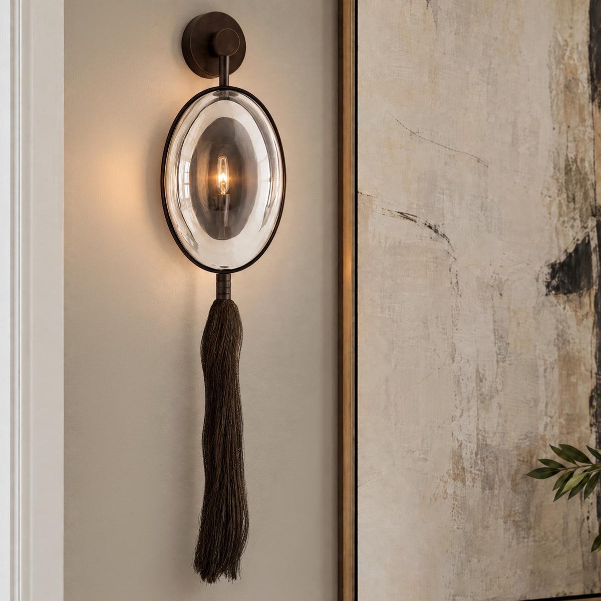 A transitional living room features the Umbria Wall Sconce 37", Bronze with an oval glass shade and dark tassel mounted beside framed abstract artwork.