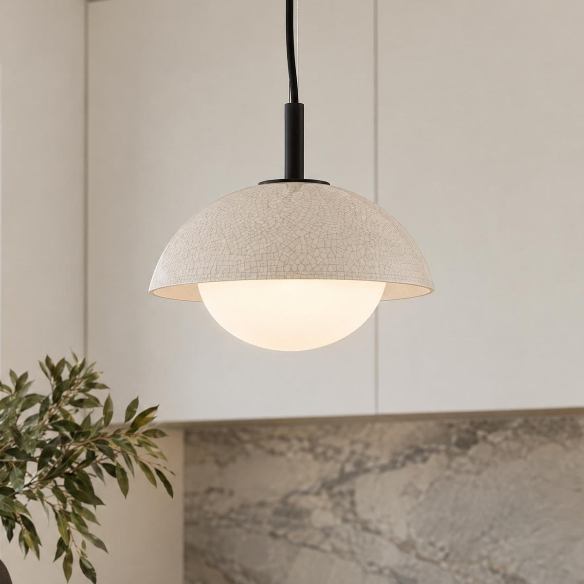 A modern kitchen featuring a matte black and ivory crackle ceramic pendant light suspended above a vein-patterned stone backsplash and off-white cabinets.